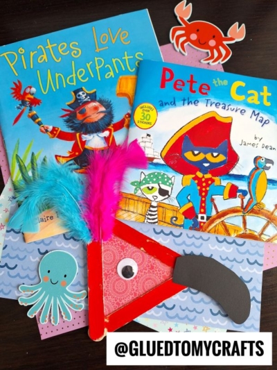 Two colorful pirate-themed children’s books, Pirates Love Underpants and Pete the Cat and the Treasure Map, are displayed with pirate and sea creature crafts, including a red pirate hat, a googly eye, and a vibrant craft stick parrot.
