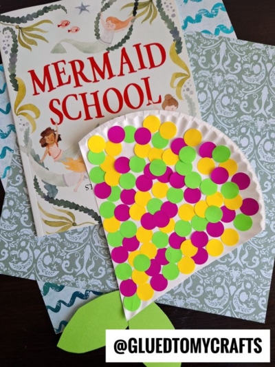 A children’s book titled “Mermaid School” sits next to a paper plate mermaid tail craft shaped like a fish, decorated with bright yellow, pink, and green dots for a fun polka dot mermaid theme. Patterned papers and @GLUEDTOMYCRAFTS are also visible.