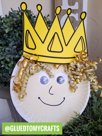 A paper plate princess craft featuring a smiling face with googly eyes, a large yellow paper crown, and curly gold ribbon hair, placed in front of green plants. @GLUEDTOMYCRAFTS is written in a green box at the bottom.