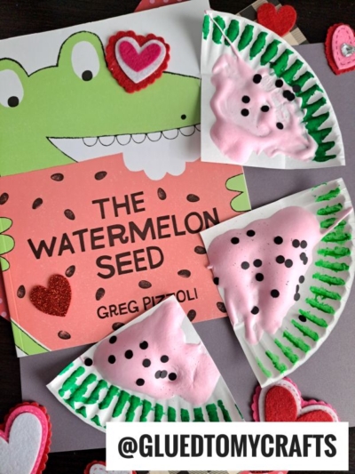 A children’s book titled The Watermelon Seed is surrounded by heart decorations and three puffy paint watermelon crafts—paper plates painted pink with puffy paint and dotted with black seeds for a fun, creative look.