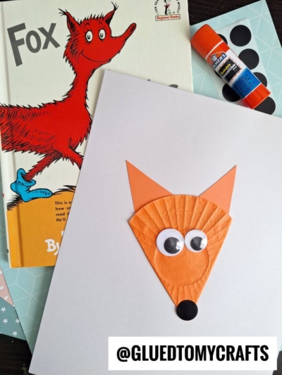 A fox-themed children’s book, a glue stick, colored paper, and a cupcake liner fox craft with googly eyes and paper ears are arranged on a table. Perfect for making cupcake liner fox cards! @GLUETOMYCRAFTS is written at the bottom.