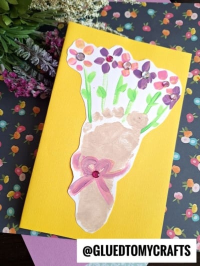 A handmade footprint flower bouquet card with a painted footprint as colorful blooms, tied with a pink ribbon. The yellow card is set against floral papers and real flowers. Text reads @GLUEDTOMYCRAFTS.