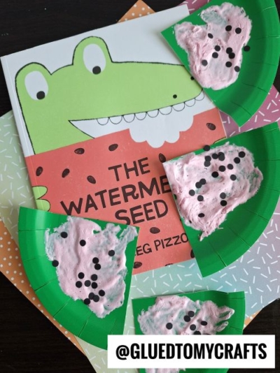 A children’s book titled The Watermelon Seed with a cartoon crocodile on the cover is surrounded by green paper plate puffy paint watermelon crafts, featuring pink puffy paint and black dots to resemble seeds.