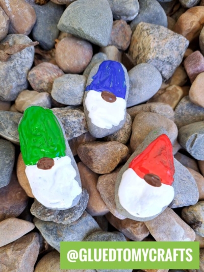 Three painted rocks designed as gnome faces with red, green, and blue hats and white beards sit on a bed of pebbles. A green label at the bottom reads @GLUEDTOMYCRAFTS.