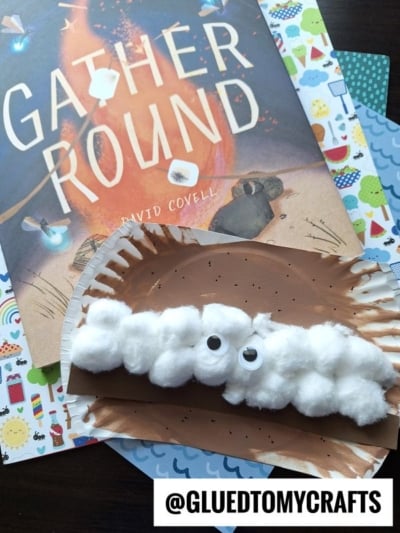 A children’s craft project features a half-paper-plate painted brown with cotton balls and two googly eyes, resembling a sheep—much like a playful paper plate smores creation—placed on colorful paper next to the book Gather Round by David Covell.