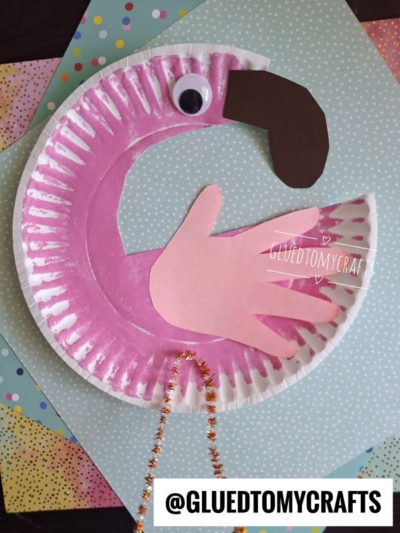 A paper plate flamingo craft featuring a pink paper hand wing, a googly eye, black paper beak, and sparkly orange pipe cleaner legs—all set on pastel polka dot papers. This cute paper flamingo is perfect for creative fun!.