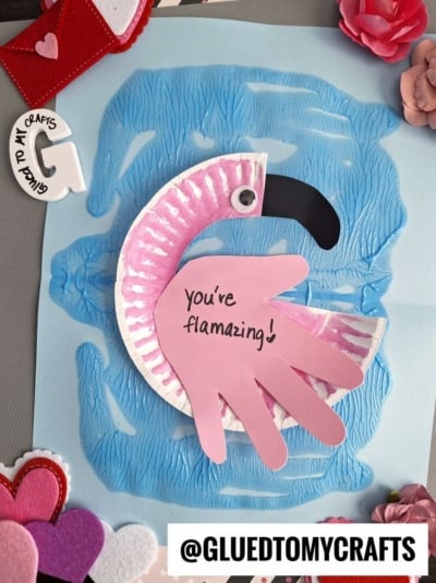 A paper plate flamingo craft made from a handprint sits on blue textured paper, surrounded by felt and paper hearts. “You’re flamazing!” is written on the handprint. @GLUEDTOMYCRAFTS is at the bottom.