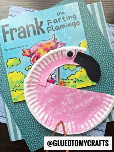 A children's book titled Frank the Farting Flamingo lies under a paper plate flamingo craft, complete with a googly eye and beak, on blue patterned paper. @GLUEDTOMYCRAFTS is labeled at the bottom.