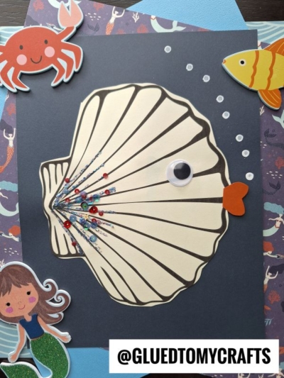 A paper seashell fish with a googly eye, heart-shaped mouth, glitter, and sequins to look like a fish is surrounded by cartoon sea creature stickers on a dark background. @GLUEDTOMYCRAFTS is labeled at the bottom.