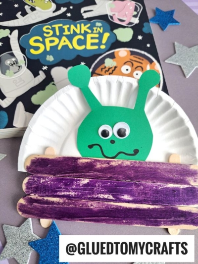 A children’s craft features a green alien with googly eyes made from paper and a paper plate, sitting in a popsicle stick alien spaceship painted purple. A book titled Stink in Space! sits in the background, along with silver star cutouts.