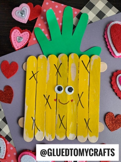 A popsicle stick pineapple craft for kids made from yellow-painted sticks, a green paper hand cutout for leaves, googly eyes, and a red smile, surrounded by red and pink felt hearts on colored paper.