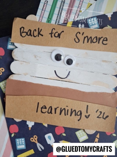A popsicle stick smore craft shaped like a s’more, complete with googly eyes and the words "Back for Smore learning! 26" written on it, lying on a colorful, school-themed background. This adorable popsicle stick smores project is perfect for welcoming students back to the classroom!.