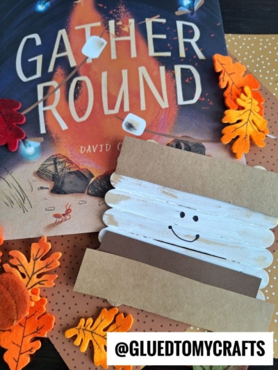 A children’s book titled “Gather Round” is surrounded by felt autumn leaves, dotted paper, and a popsicle stick smores craft with a smiling face. The handle @GLUEDTOMYCRAFTS appears at the bottom.
