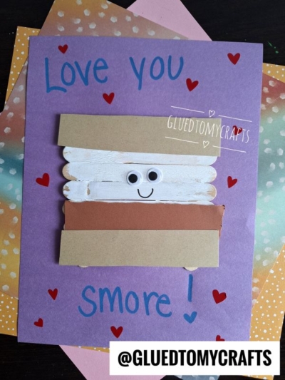 A purple card with Love you Smore! written in blue, decorated with red hearts. A popsicle stick smore made from craft sticks, paper, and googly eyes sits in the center. The handle @GLUEDTOMYCRAFTS is at the bottom.
