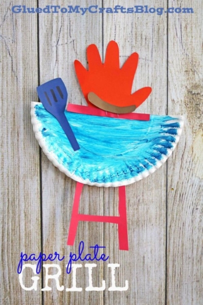 This creative paper plate grill craft resembles a real grill, featuring a painted blue top, a red handprint as a flame, and sturdy red paper legs. A blue spatula shape is attached to the left side of the plate for added detail. The background is wood, enhancing the cookout-inspired theme.