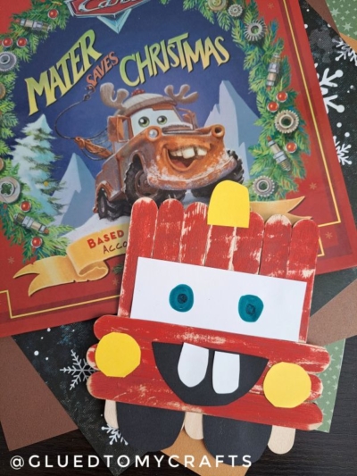 A popsicle stick Mater kid craft idea, painted red with paper details, sits atop a “Mater Saves Christmas” book cover featuring Mater with festive reindeer antlers.