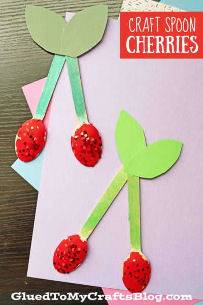 Two cherry crafts made from craft spoon cherries and colored paper sit on pastel sheets. The cherries feature red, glittery tops and green leaves, with "Craft Spoon Cherries" in the top right corner.