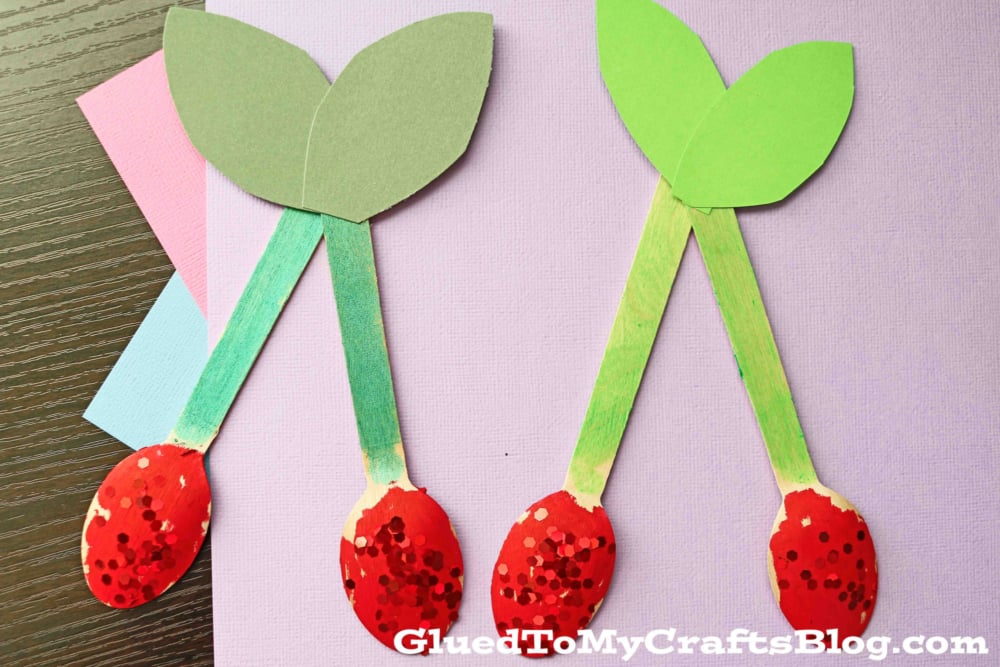 Two cherry crafts made from green construction paper stems and leaves, with red, oval-shaped craft spoon cherries as tops, are placed on light purple paper. A watermark reads GluedToMyCraftsBlog.com.