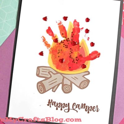A handmade card featuring a painted handprint campfire with logs and vibrant flames, decorated with small red dots. The card reads Happy Camper and is surrounded by colorful paper, perfect for adventurous spirits.