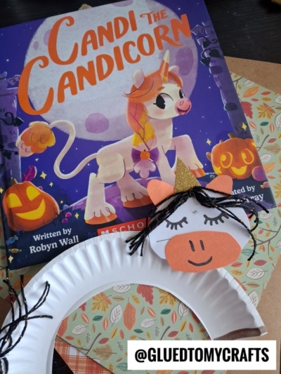 A children’s book titled Candi the Candicorn features a unicorn with a candy corn horn, surrounded by pumpkins. In front is a paper plate unicorn craft with black yarn hair, gold horn, and orange snout. A tag reads @GLUEDTOMYCRAFTS.