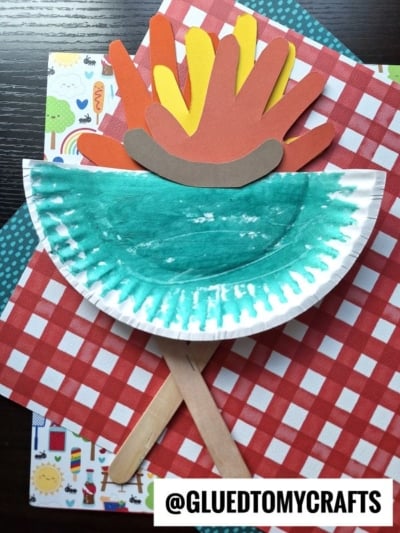 A paper plate grill painted green is folded in half on top of red checkered and patterned paper. Above it is a craft campfire made from colored paper hands and sticks. @GLUEDTOMYCRAFTS is at the bottom right.