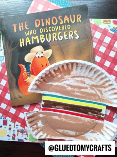 A children’s book titled The Dinosaur Who Discovered Hamburgers sits next to a paper plate hamburger craft, all atop a red checkered tablecloth. The handle @GLUEDTOMYCRAFTS is at the bottom.