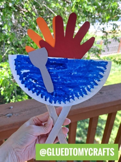 A hand holds a paper plate grill craft, painted to resemble water, with two paper handprints as a sunset and a gray paper spatula, set against trees and a wooden fence. @GLUEDTOMYCRAFTS is written at the bottom.