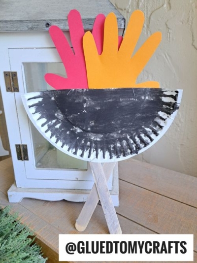 A paper plate grill painted black and folded in half forms a cauldron, with red and orange handprints as flames and two craft sticks as logs, displayed in front of a white lantern.