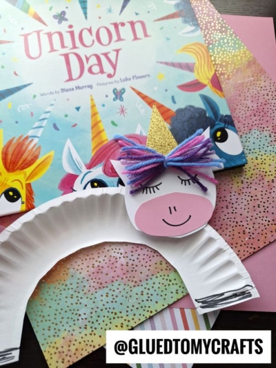 A whimsical paper plate unicorn craft with yarn hair and a gold horn sits atop the Unicorn Day book, surrounded by pink paper and vibrant backgrounds. Text reads @GLUEDTOMYCRAFTS at the bottom.