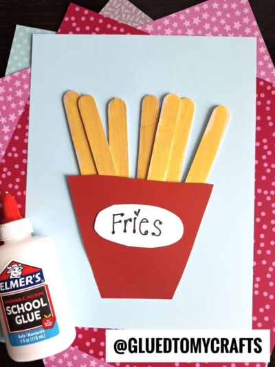 A craft project featuring popsicle stick fries painted yellow, arranged in a red paper container labeled "Fries," with a bottle of Elmer's school glue and colorful patterned paper in the background.