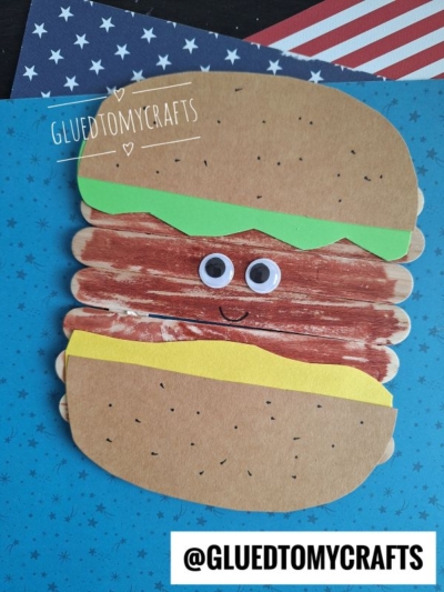 A hamburger kid craft made from colored paper and popsicle sticks with googly eyes and a smile sits on a blue background with stars. The top reads “GLUEDTOMYCRAFTS” and the bottom says “@GLUEDTOMYCRAFTS”.