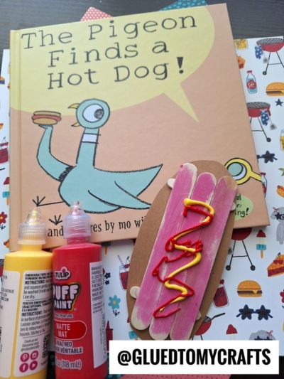 A children’s book titled The Pigeon Finds a Hot Dog! is on a table with craft materials, including a craft stick hot dog, yellow and red paint, and a printed Instagram handle @GLUEDTOMYCRAFTS.