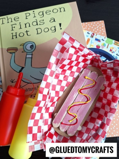A children’s book titled The Pigeon Finds a Hot Dog! sits next to a popsicle stick hot dog decorated with pink and yellow accents, flanked by red and yellow squeeze bottles and colorful paper in the background.