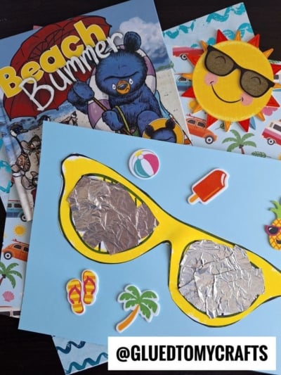 Colorful beach-themed crafts, including a sunglasses kid craft with a large yellow mixed media sunglasses cutout, a smiling sun with shades, and beachy stickers, all displayed on top of a Beach Bummer book. @GLUEDTOMYCRAFTS at the bottom.