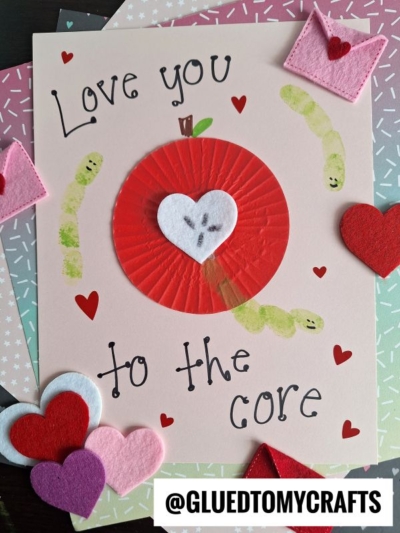 A handmade cupcake liner apple card with the phrase "Love you to the core," featuring a red cupcake liner apple with a white heart center, felt hearts, paper worms, and charming red heart accents.