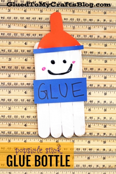 A popsicle stick glue bottle craft, painted white with a blue GLUE label, red cap, and a smiling face, rests on a playful background of overlapping wooden rulers.