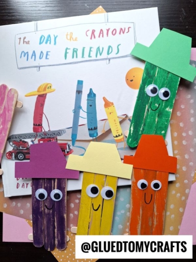 Four popsicle stick crayon friends with smiling faces and colorful paper hats are displayed on a children’s book titled The Day the Crayons Made Friends. This fun popsicle stick crayon craft for kids uses green, yellow, purple, and orange crayons.