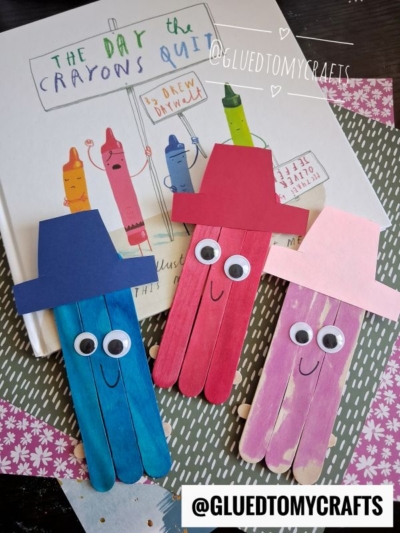 Three colorful popsicle stick crayon friends with hats and googly eyes are placed on a copy of “The Day the Crayons Quit” book. Each character is red, purple, or blue with different colored hats, making this a fun popsicle stick crayon craft for kids.