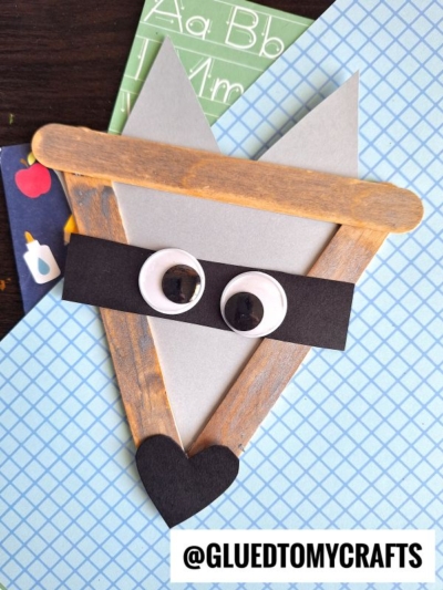 A Popsicle Stick Raccoon face made from craft sticks, gray paper, a black paper mask, large googly eyes, and a black heart-shaped nose, displayed on a blue grid background with @GLUEDTOMYCRAFTS label.