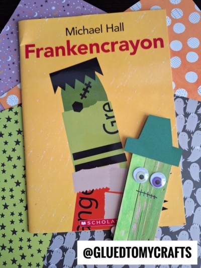 A yellow Frankencrayon book lies on Halloween-themed papers, accompanied by a green Frankenstein craft made from a painted popsicle stick, googly eyes, and a paper hat—an adorable popsicle stick crayon craft for kids. The @gluedtomycrafts label appears at the bottom.