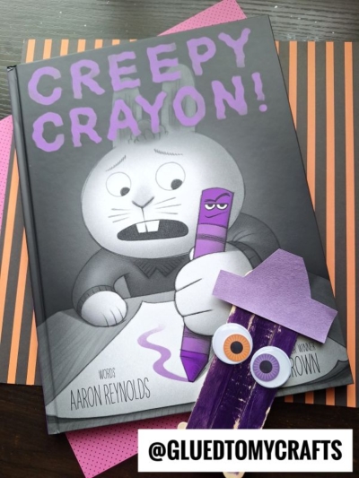 The book Creepy Crayon! shows a rabbit holding a worried-looking purple crayon. Beside it is a fun popsicle stick crayon craft for kids, featuring googly eyes and a frown to match the story’s theme. Striped and polka dot paper is underneath, making it perfect inspiration for your own popsicle stick crayon friends. @gluedtomycrafts.