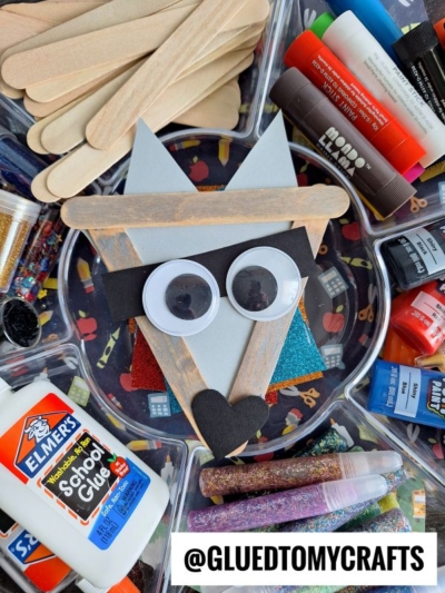 A wolf craft made from popsicle sticks, googly eyes, and paper sits on a tray surrounded by glue, glitter, markers, and other craft supplies—perfect for inspiring your next Popsicle Stick Raccoon project. A label reads @GLUEDTOMYCRAFTS.