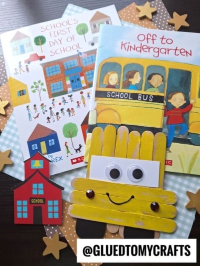Two children’s books about starting school, a popsicle stick school bus craft with googly eyes, a red paper schoolhouse, and gold star decorations are arranged on a table.