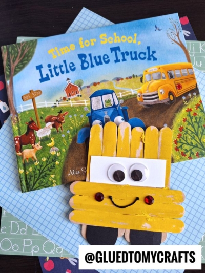 A children's book titled Time for School, Little Blue Truck sits behind a cheerful popsicle stick school bus craft with googly eyes and a smile. Blue and alphabet-patterned paper decorate the surface.