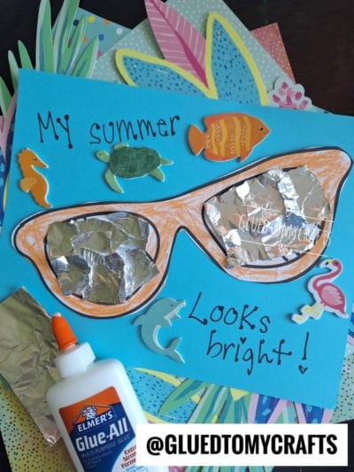 A colorful summer sunglasses kid craft features orange mixed media sunglasses made of foil on blue paper, decorated with sea animal stickers, handwritten “My summer looks bright!”, and a bottle of Elmer’s glue.