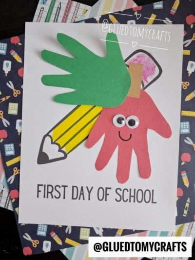 Handprint paper art of a pencil and two hands, one green and one red with googly eyes and a smile, on a card reading FIRST DAY OF SCHOOL. This adorable handprint keepsake makes the perfect first day of school keepsake for capturing special memories. The background features playful school supply patterns. @GLUEDTOMYCRAFTS is written twice.