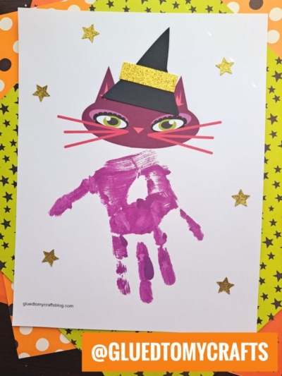 A Halloween craft features a handprint Halloween cat with a black hat, its body made from a purple handprint, surrounded by gold star stickers on white paper, with an orange tag that reads @GLUEDTOMYCRAFTS.