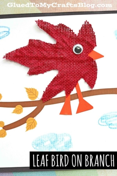 A red burlap leaf shaped like a bird, featuring a googly eye and orange paper beak and legs, is perched on a drawn branch with leaves against white paper with blue clouds—a charming bird craft idea perfect for creating your own whimsical leaf bird on branch scene.