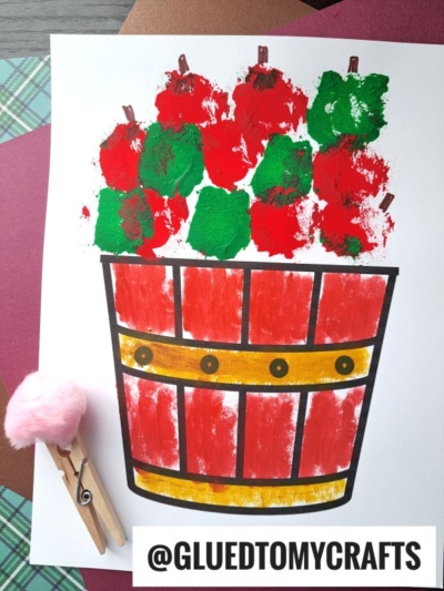 A child’s craft of a bucket filled with red and green pom pom stamped apples, made with a clothespin and pom-pom stamper, is shown on colored paper. The username @GLUEDTOMYCRAFTS appears at the bottom.