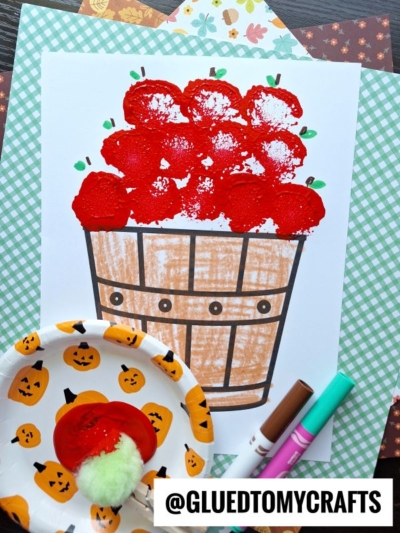 A kids craft showing a basket filled with red apples, created with paint and markers. A small plate with pumpkin designs holds red paint and a pompom, suggesting a fun pom pom stamped apples technique.